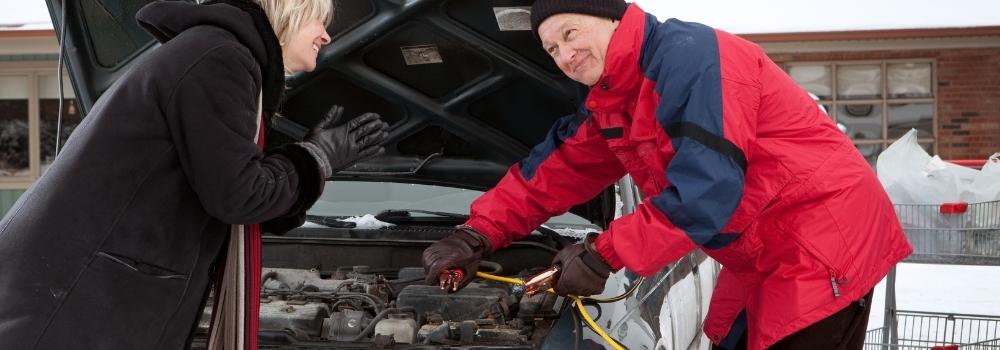 An older man in a winter coat showing his neighbour how to boost a battery.