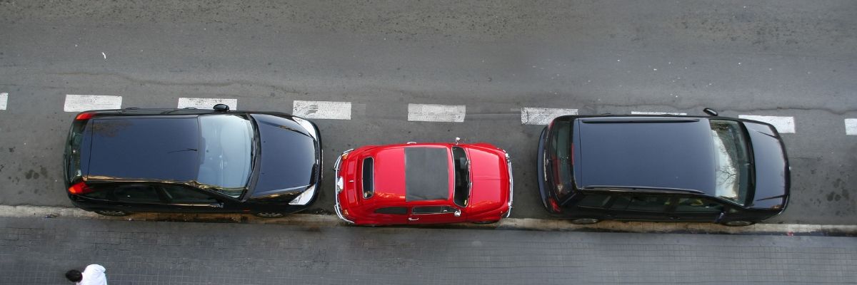 A small vs. large car comparison between two large black SUVs and a small red compact sedan in between them parked on the side of the road.