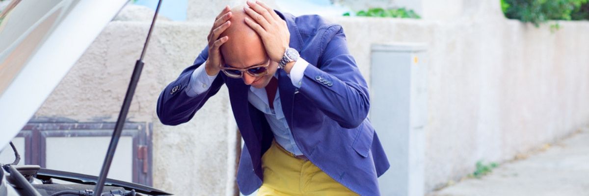 A bald man in a purple jacket and yellow pants holding his head looking at his car's engine while dialling a claim for car insuranc in Ontario.