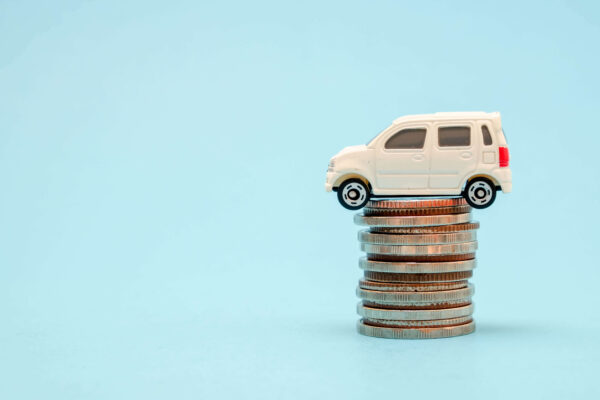 A toy white vehicle sitting on top of a stack of coins. The tiny driver is wondering: "Why is My Car Insurance Deductible so High?"